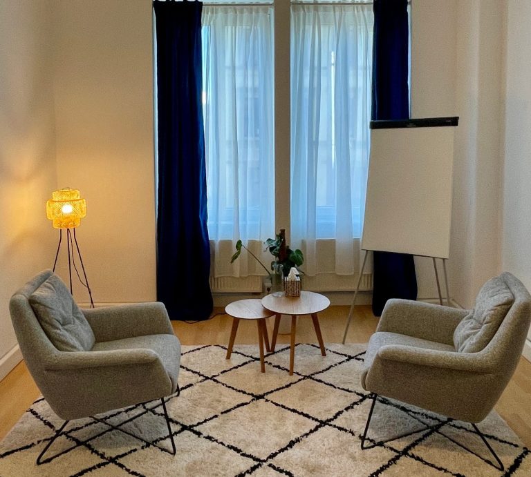 Gemütlicher Therapieraum mit zwei Sesseln, Tisch, Pflanze und Flipchart vor großen Fenstern mit weißen und blauen Vorhängen.