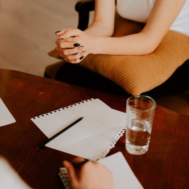Hände mit schwarzen Nägeln auf einem Tisch mit Notizblock und Glas Wasser.