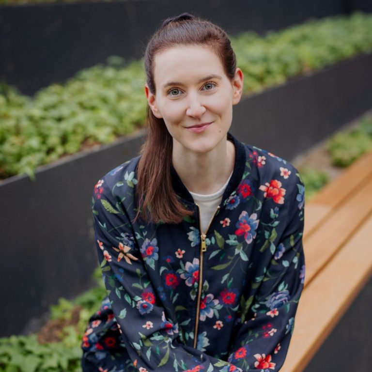 Foto der Therapeutin: Junge Frau mit braunen Haaren  Blumenmuster-Jacke sitzt auf einer Bank vor grünen Pflanzen und lächelt.