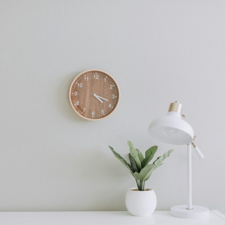Holzwanduhr, moderne Tischlampe und eine kleine Grünpflanze in einem weißen Pflanzentopf in einem hellen Raum.